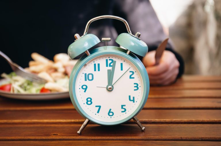 Close-up of an alarm clock with a blurred background of various healthy and nutritious foods, symbolizing the importance of timely and mindful dietary choices for overall well-being and longevity as advised by Dr. Peter Attia.