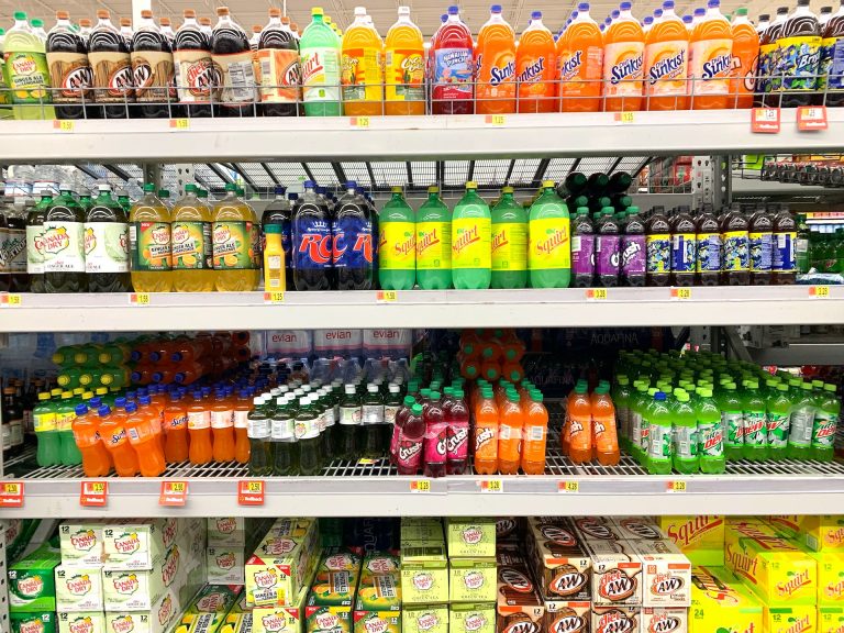 Grocery store shelves packed with various brands of soda, a prominent source of fructose, alongside a section dedicated to cooking oils, including pro-inflammatory seed oils, both hidden contributors to the global obesity epidemic.